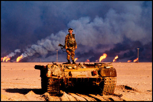 An American soldier stands on top of a destroyed Iraqi tank. Kuwaiti oil wells, ignited by Saddam's retreating forces, burn in the distance, Kuwait