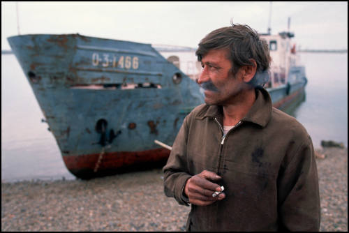 A Siberian fisherman smokes next to his fishing vessel on the shore of the Tom River, Tomsk, Russia